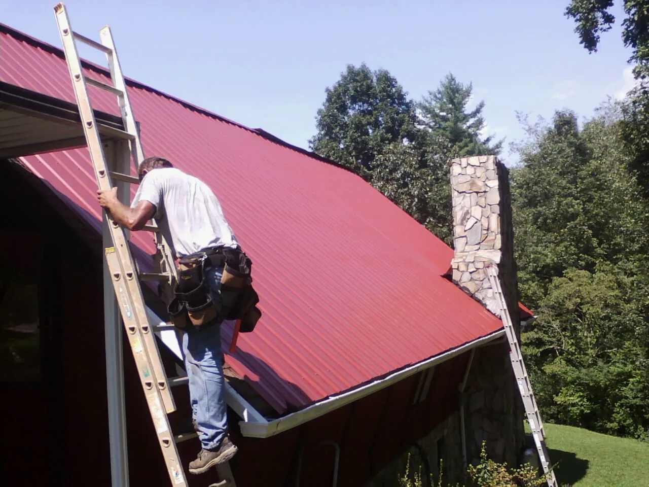 Roofer on a ladder beside a red metal roof and stone chimney — Soffit & Fascia Repair by Roofing Brothers in Big Bend